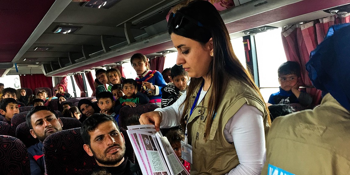 Eine Frau steht in einem vollen Bus und verteilt Flyer auf denen Minenaufklärung zu sehen ist.